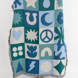 A patchwork throw blanket with geometric and folk patterns in blue, green, and off-white hues, featuring a fringe border.